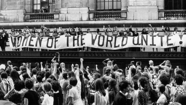 Mujeres reunidas ante un cartel que dice, "¡Mujeres del mundo unidas!" en inglés